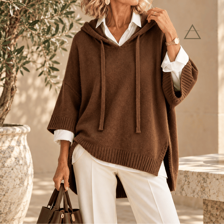 Poncho Clémence en maille chocolat, à capuche et finitions côtelées, porté sur une chemise blanche avec un pantalon blanc.
