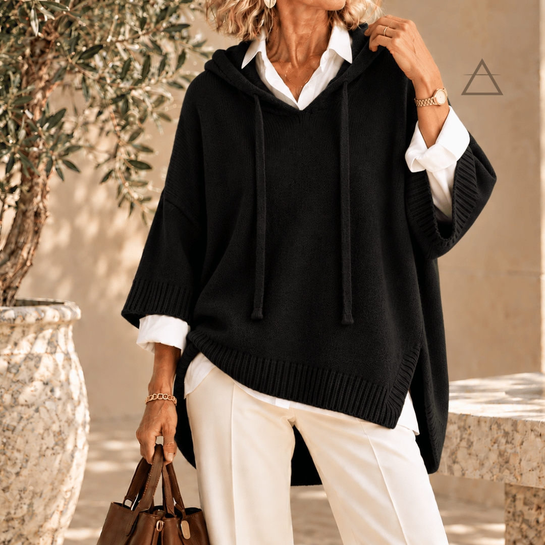 Poncho Clémence en maille noir, à capuche et finitions côtelées, porté sur une chemise blanche avec un pantalon blanc.

