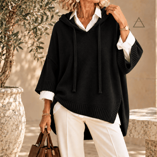 Poncho Clémence en maille noir, à capuche et finitions côtelées, porté sur une chemise blanche avec un pantalon blanc.
