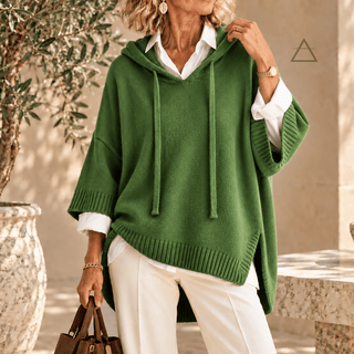 Poncho Clémence en maille vert prairie, à capuche et finitions côtelées, porté sur une chemise blanche avec un pantalon blanc.

