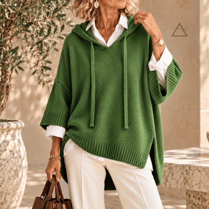 Poncho Clémence en maille vert prairie, à capuche et finitions côtelées, porté sur une chemise blanche avec un pantalon blanc.

