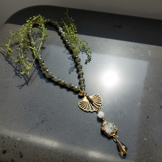 Collier Labradorite Moonlight aux perles gris fumé, avec éléments dorés et pendentif en quartz, présenté sur un fond sombre avec des tiges végétales.
