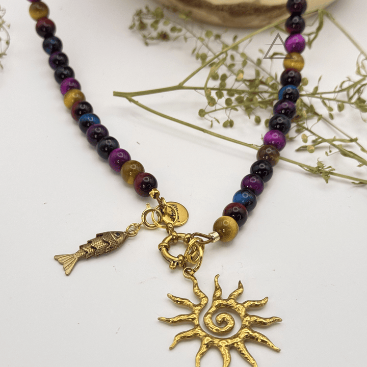 Vue rapprochée des pendentifs poisson et soleil dorés du collier SOLSTICE, avec perles multicolores sur fond blanc.