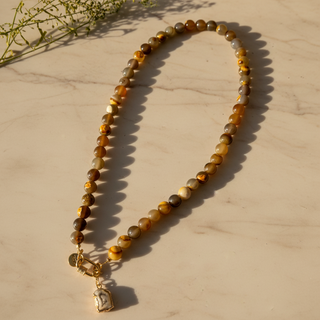 Collier Unspoken aux perles brunes, miel et grises, avec pendentif en agate, posé sur un fond clair en lumière chaude.